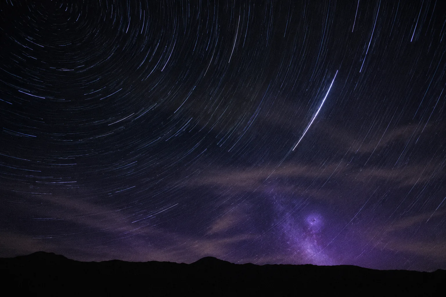 长曝光的夜空，星轨划过山脊上方的深蓝天幕，地平线附近泛着淡淡的紫色星云。